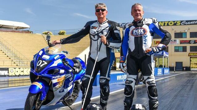 Ben en zijn coach poseren op het racecircuit. Ben heeft zijn witte stok vast, zijn andere hand rust op een blauwe motor.