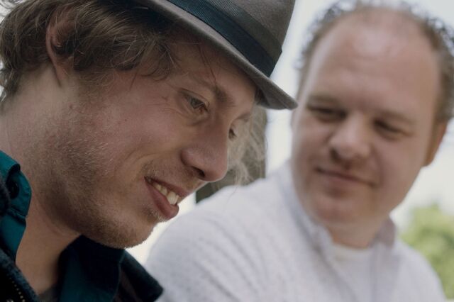 Still uit de film 80% ongeschikt. Close up van Mari Sanders, een man met bruin krulhaar. Hij heeft een hoedje op en op de achtergrond kijkt een andere man naar hem