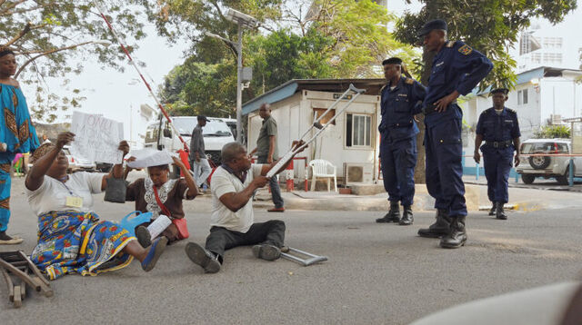 Still uit de film Downstream to Kinshasa. Enkele protestanten zitten op de grond op straat. Een van hen heft zijn kruk naar de politie