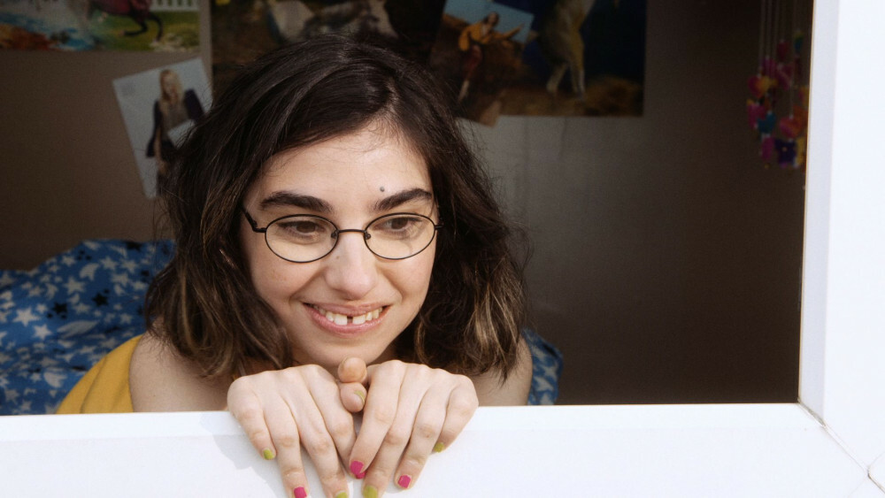 Yildiz kijkt lachend uit het raam van haar slaapkamer, haar handen rusten op de vensterbank. De muur achter haar is versierd met posters van beroemdheden.