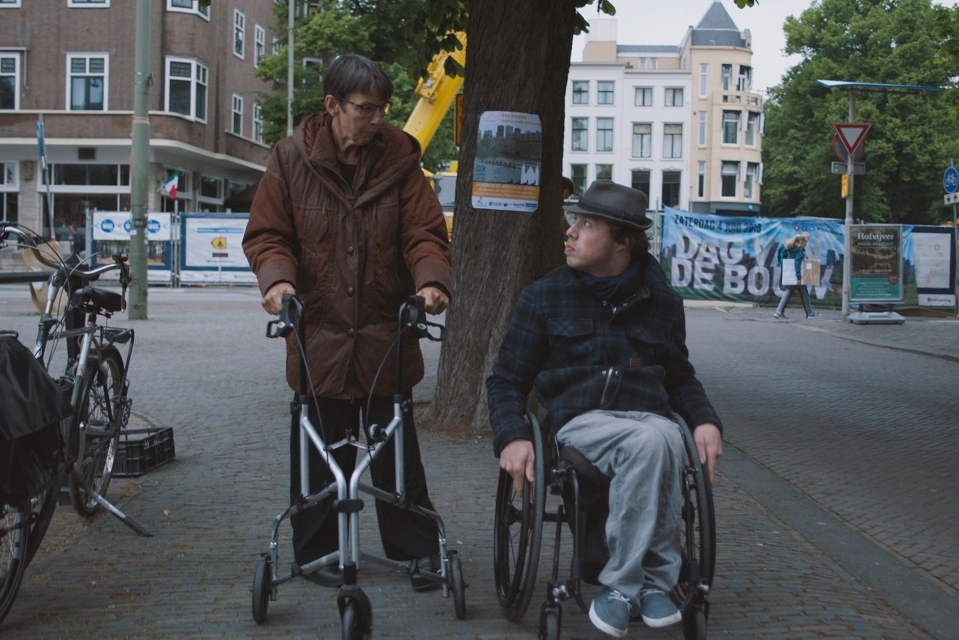 Still uit de film 80% ongeschikt. Mari Sanders, een jongeman met donker krulhaar en een hoedje, zit in een rolstoel. Naast hem loopt een vrouw met een rollator